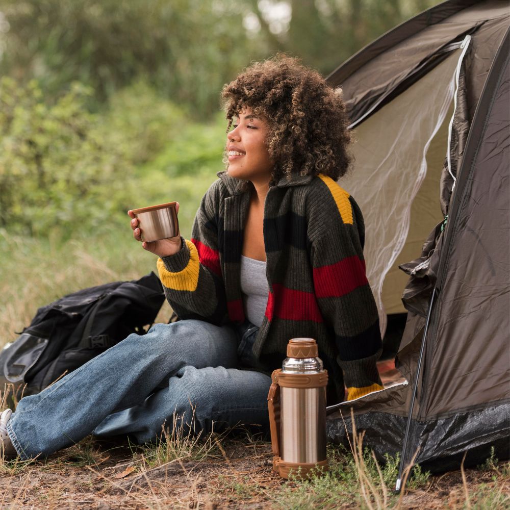 Person sitting by a tent holding a mug outdoors
