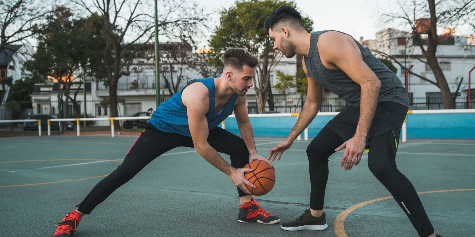 Two men playing basketball on an outdoor court with trees and buildings in the background.